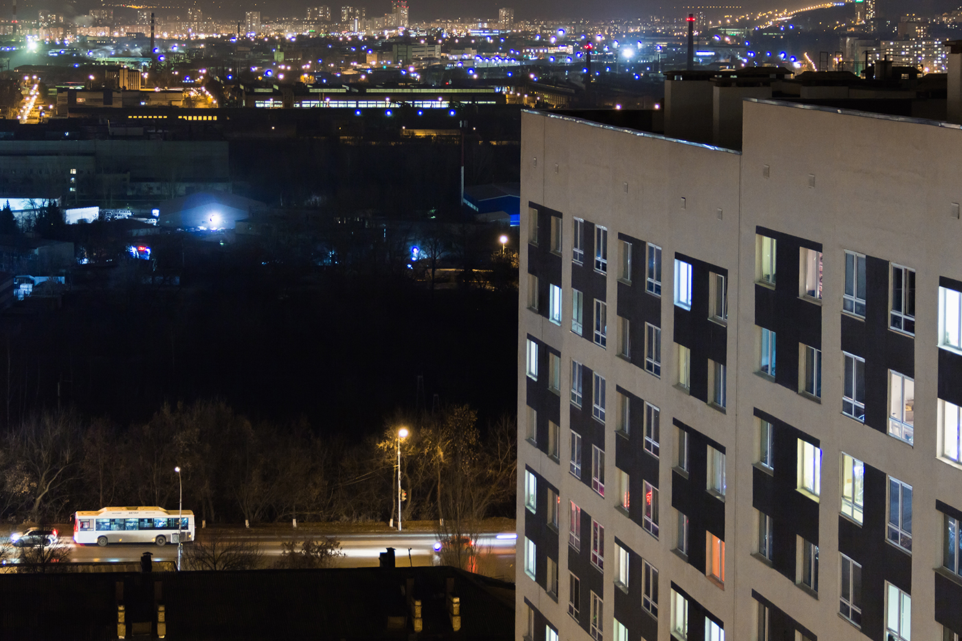 High-rise modern building in the evening window lights.Casual urban city life. Traffic on the road.From tower rooftop, industrial region with working factory and container crains.